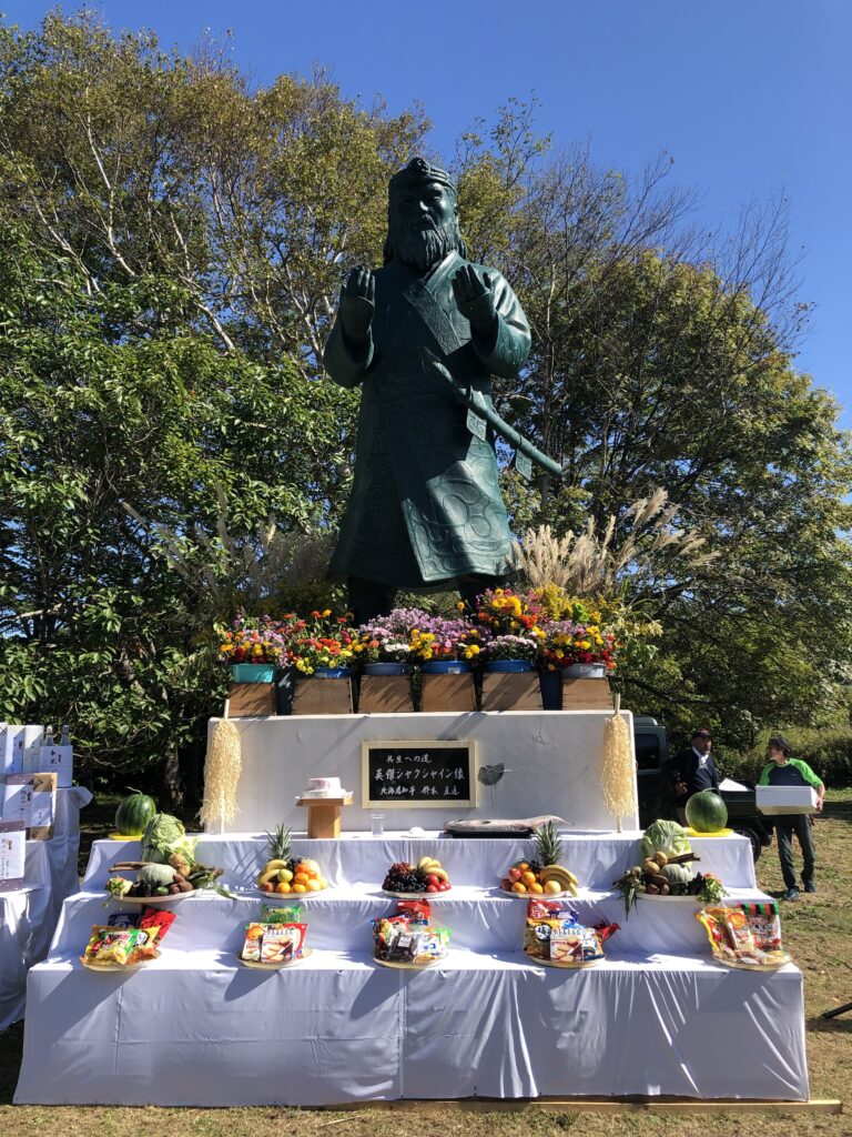 A photo of a statue of Shakushain, surrounded by memorial offerings of flowers and food.